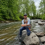 Man fishing in river, holding net, wearing blue shirt and orange vest, surrounded by rocks and green trees.
