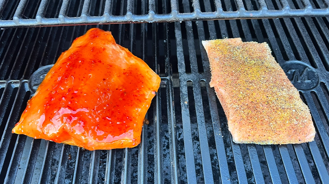 Two pieces of seasoned fish cooking on a grill, one with a red sauce and the other with a yellow seasoning.