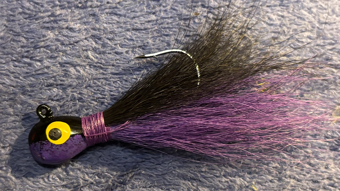 A fishing lure with a purple body, yellow eye, black and purple feathered tail, and a curved hook.